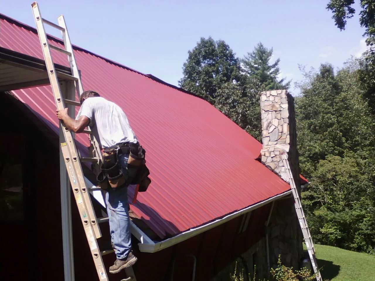 Roofer on a ladder beside a red metal roof and stone chimney — Soffit & Fascia Repair by Roofing Brothers in Inglis